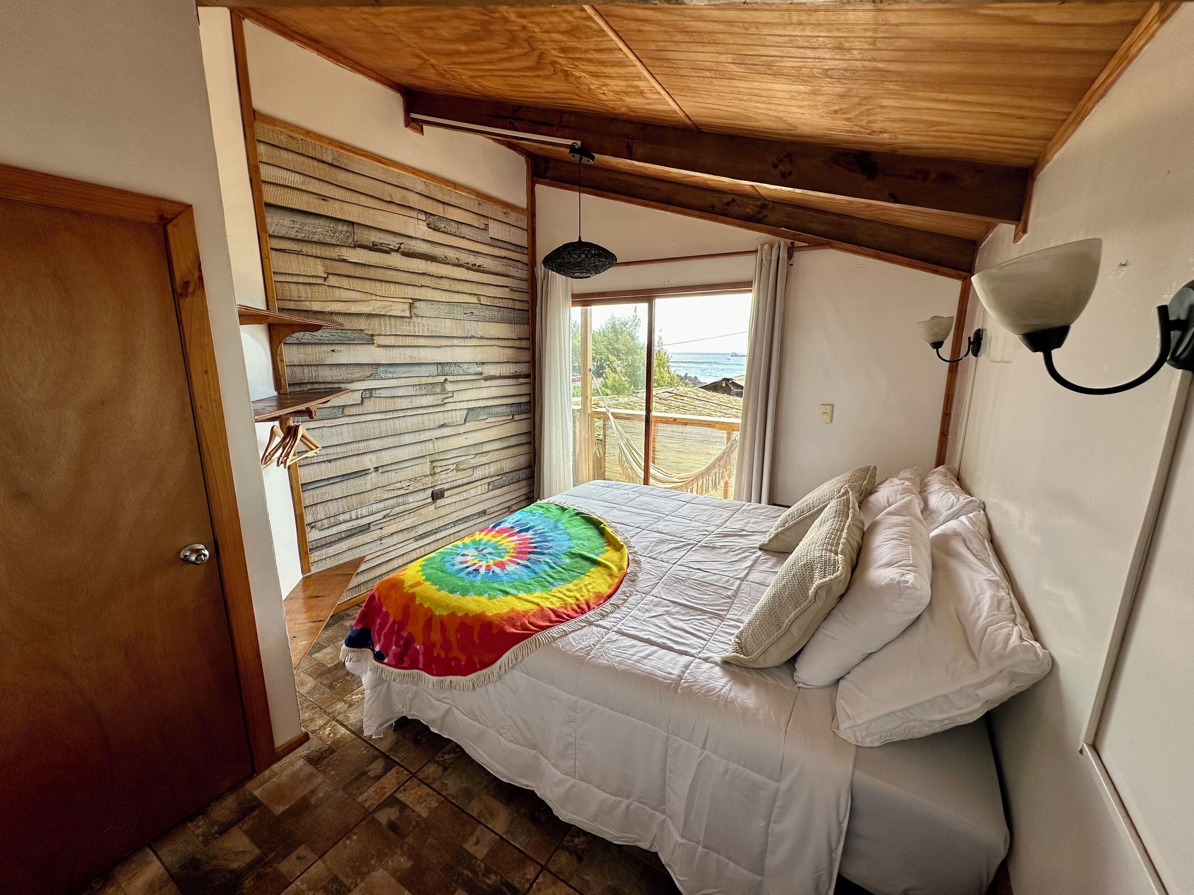 A bedroom with a wooden ceiling, a bed with a white and colorful blanket, a view of the outdoors through a window, and a wooden wall.