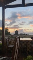 Un área de estar al aire libre con techo, con sillas de mimbre y una mesa, con vistas a una playa y una gran formación rocosa al atardecer.