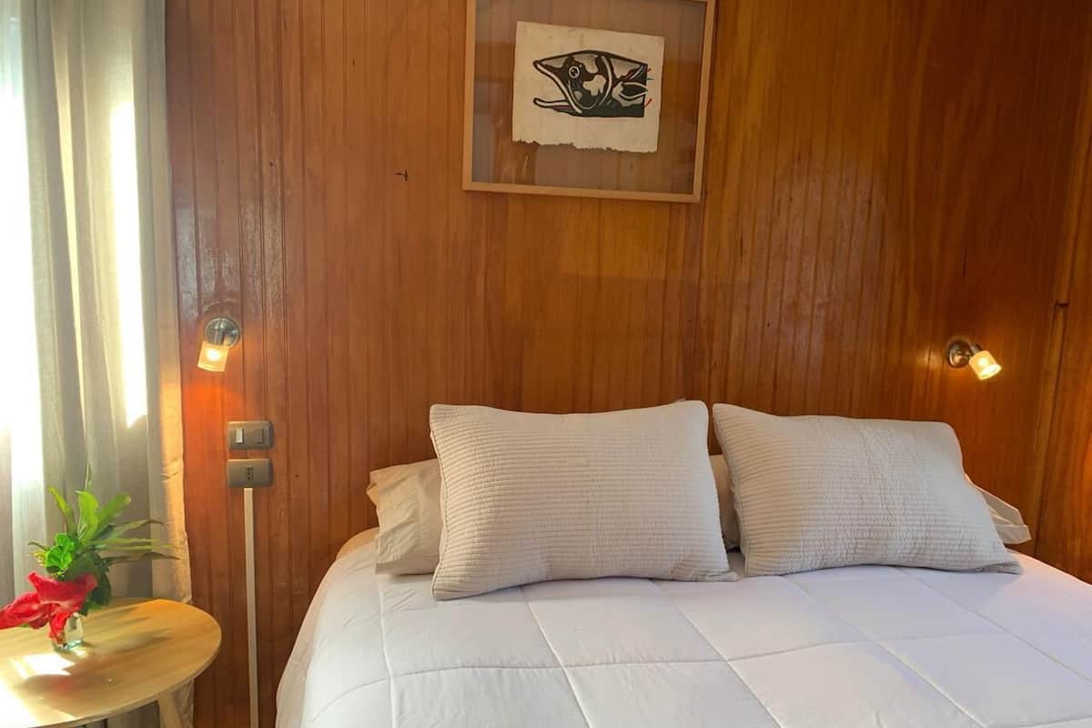 A hotel room with a wooden wall, a bed with white bedding, a small table with a plant, and a framed picture on the wall.