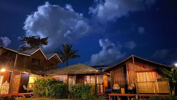 A wooden house with a thatched roof, surrounded by lush greenery and a clear night sky with clouds. - Paraíso Sunset Bungalows (Hanga Roa)