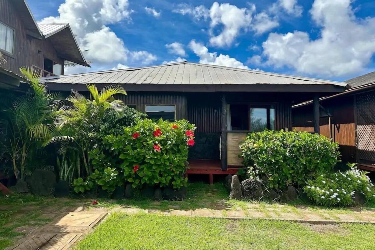 A two-story wooden house with a metal roof, surrounded by lush greenery and flowering plants.