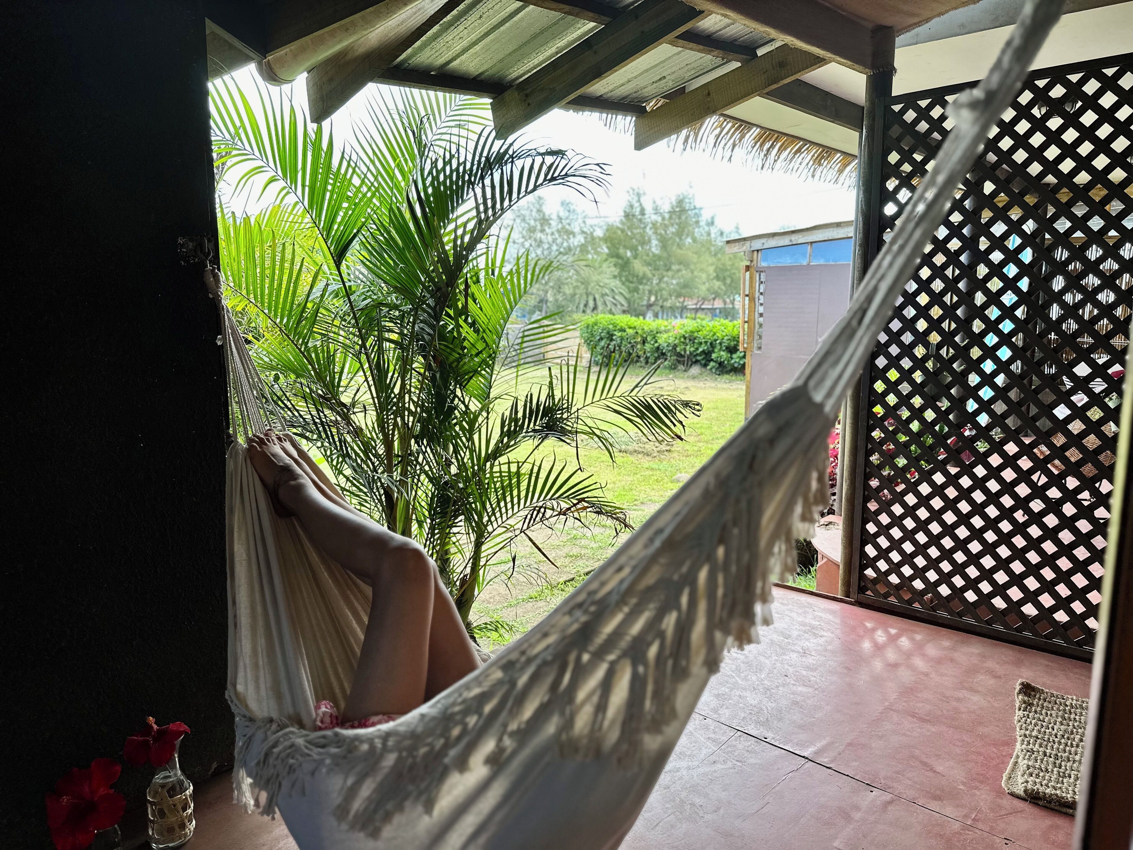 A hammock under a wooden structure with a view of a garden and a building in the background.