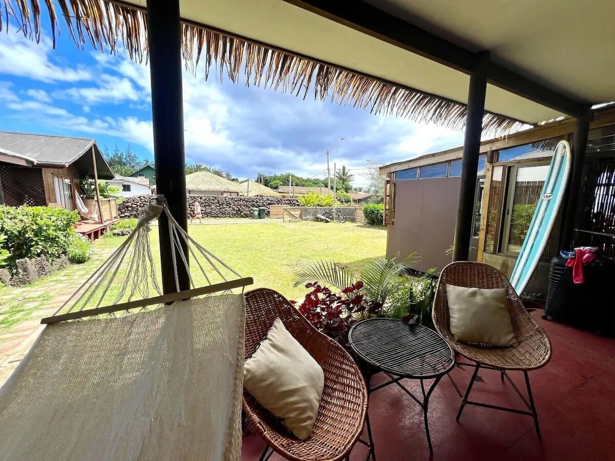 A hammock, wicker chairs, a small table, and a view of a garden and buildings.