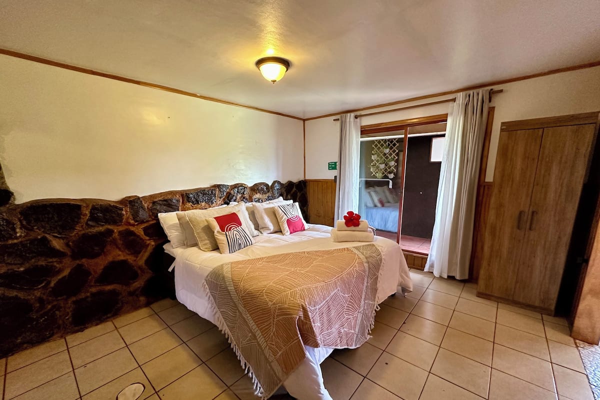 A bedroom with a stone wall, a bed with multiple pillows, a wooden headboard, a large window with curtains, and a wardrobe.