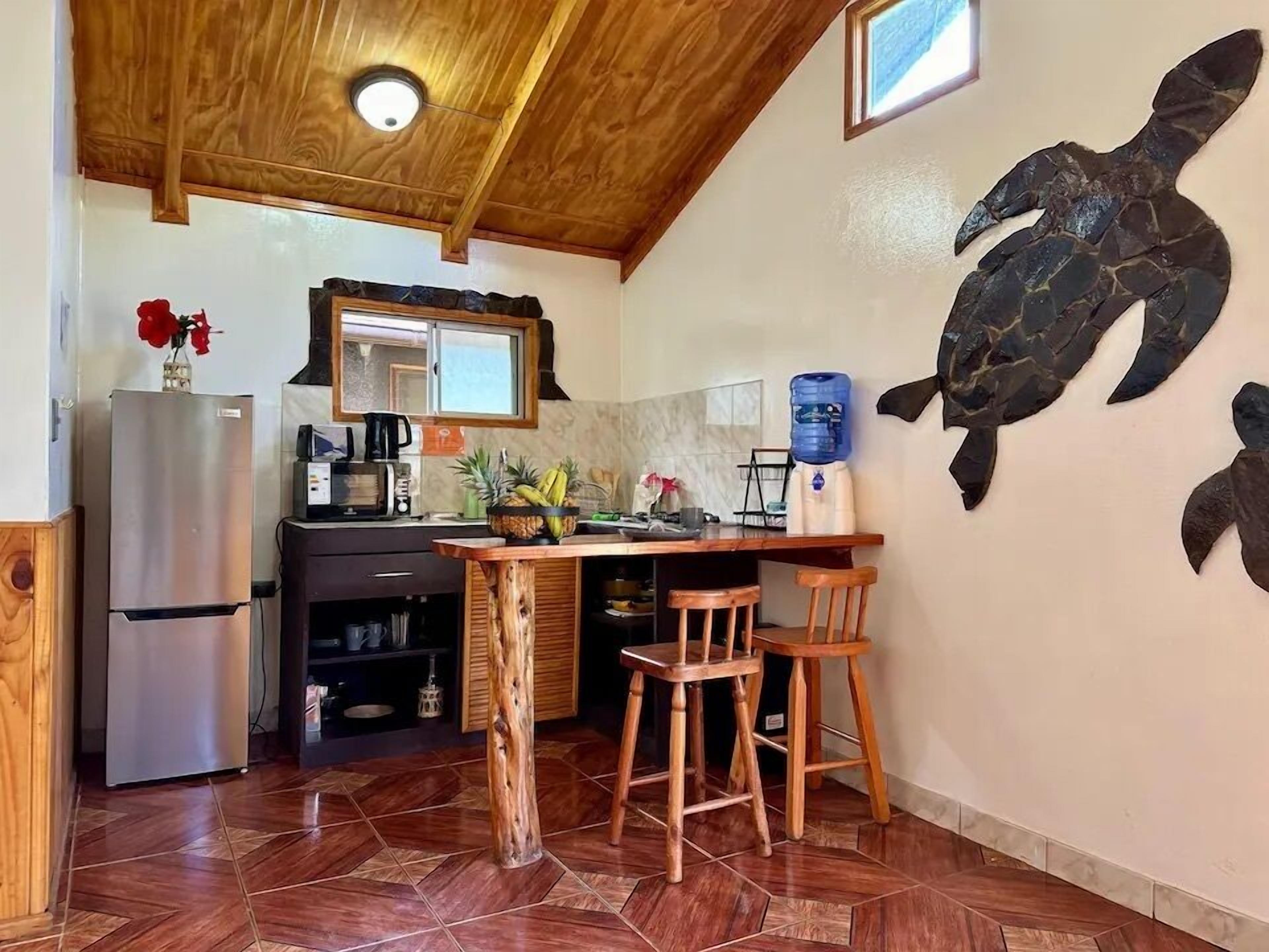 A compact kitchen with a wooden ceiling, a refrigerator, a coffee maker, and a water dispenser. There are two wooden stools at a small wooden table.