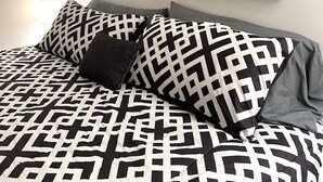 Un dormitorio con una cama que tiene una colcha de edredón con estampado geométrico en blanco y negro, una almohada en blanco y negro y una almohada gris. En la pared, encima de la cama, se exhibe una fotografía en blanco y negro de un tren sobre un puente en un área boscosa.
