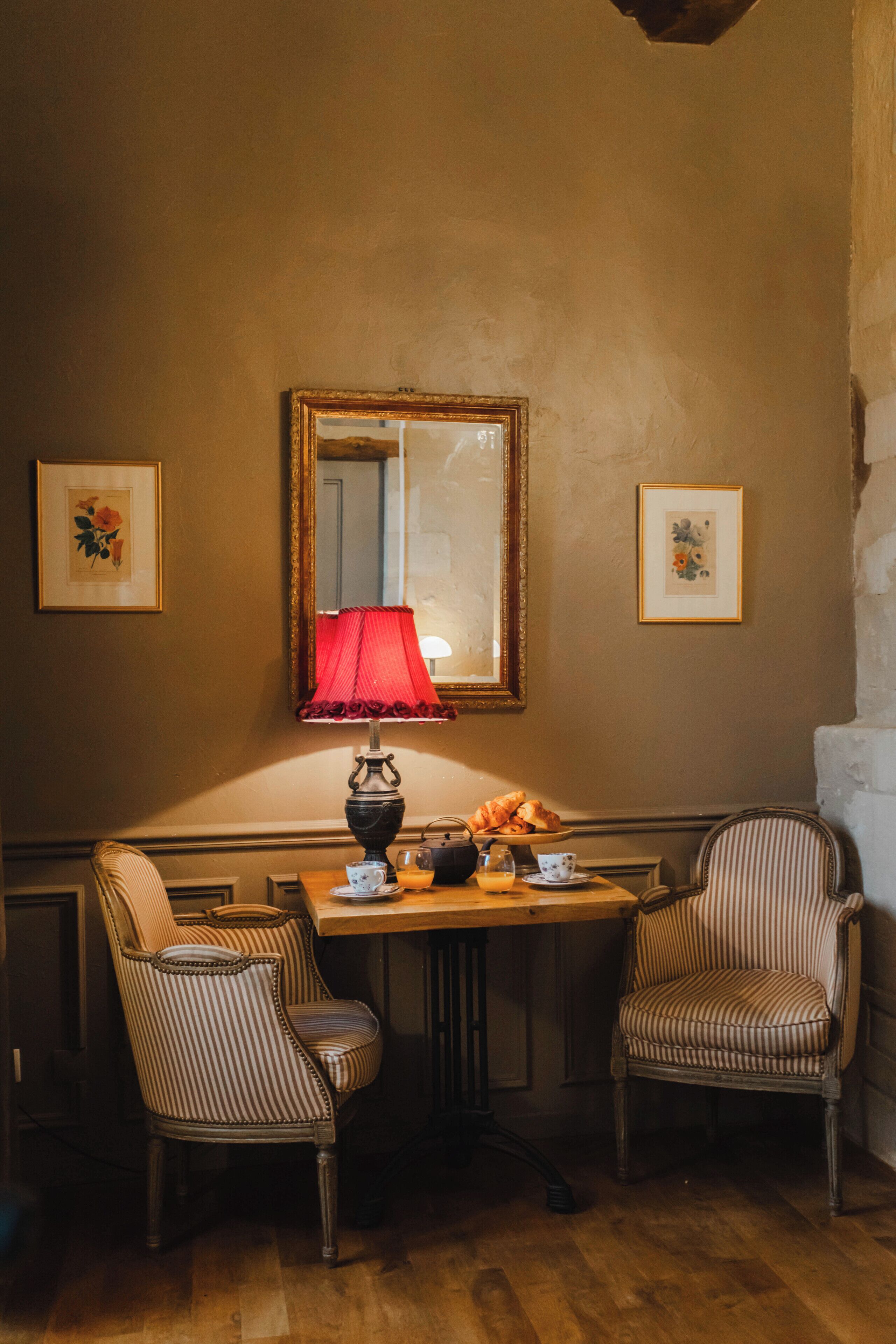 A room with a wooden table, two striped chairs, a red lamp, and framed pictures on the wall.