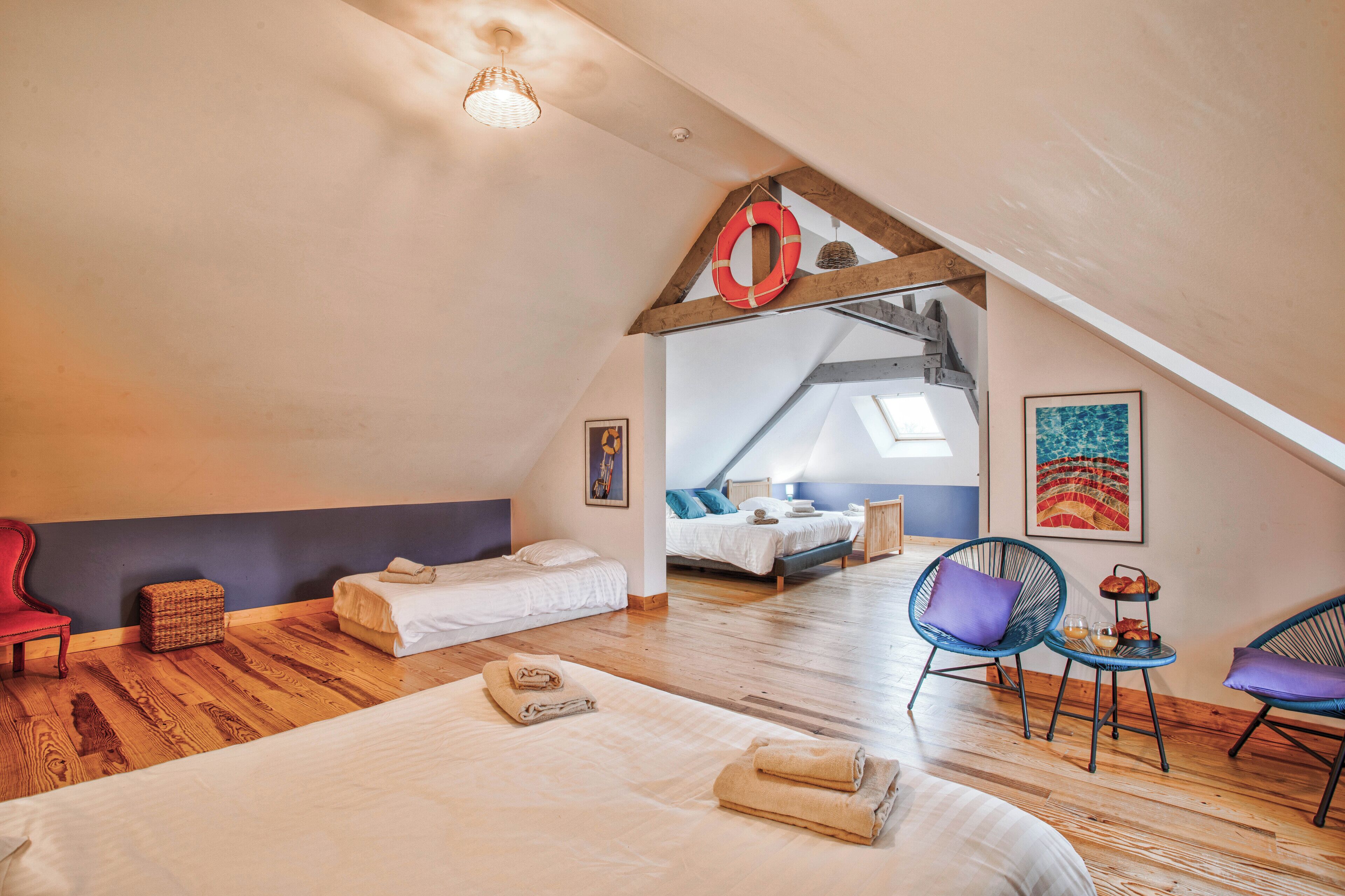A spacious attic bedroom with a sloped ceiling, a large bed, a small seating area with chairs, and a dining table with a tray of bread.