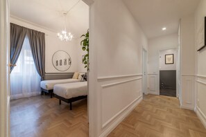 A bedroom with two beds, a chandelier, a plant, and a hallway with a door leading to another room.