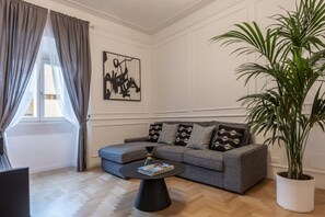 Ein Wohnzimmer mit grauem Sofa, einem schwarzen Couchtisch, einer großen Topfpflanze und einem Fenster mit grauen Vorhängen.
