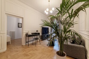 A living room with a wooden floor, a grey sofa, a black chair, a desk with a computer, and a large potted plant.
