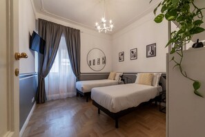A bedroom with two single beds, a TV mounted on the wall, a chandelier, and a plant.