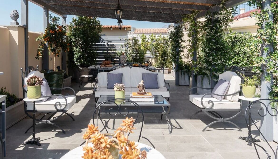 An outdoor patio with a sofa, chairs, and a table, surrounded by potted plants and a pergola.