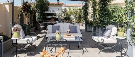 Un patio exterior con un sofá, sillas y una mesa, rodeado de plantas en macetas y una pérgola.