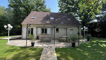 A house with a tiled roof, a covered patio with a table and benches, and two lamp posts.