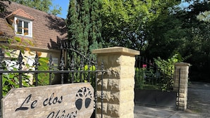 A wooden sign with the text "Le dos des Cheves" mounted on a stone pillar, with a metal gate and a house in the background.
