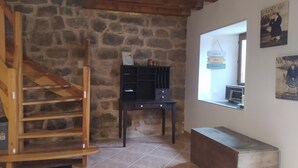 Un interior rústico con una escalera de madera, paredes de piedra, una pequeña mesa negra y un ventanal con una obra de arte.