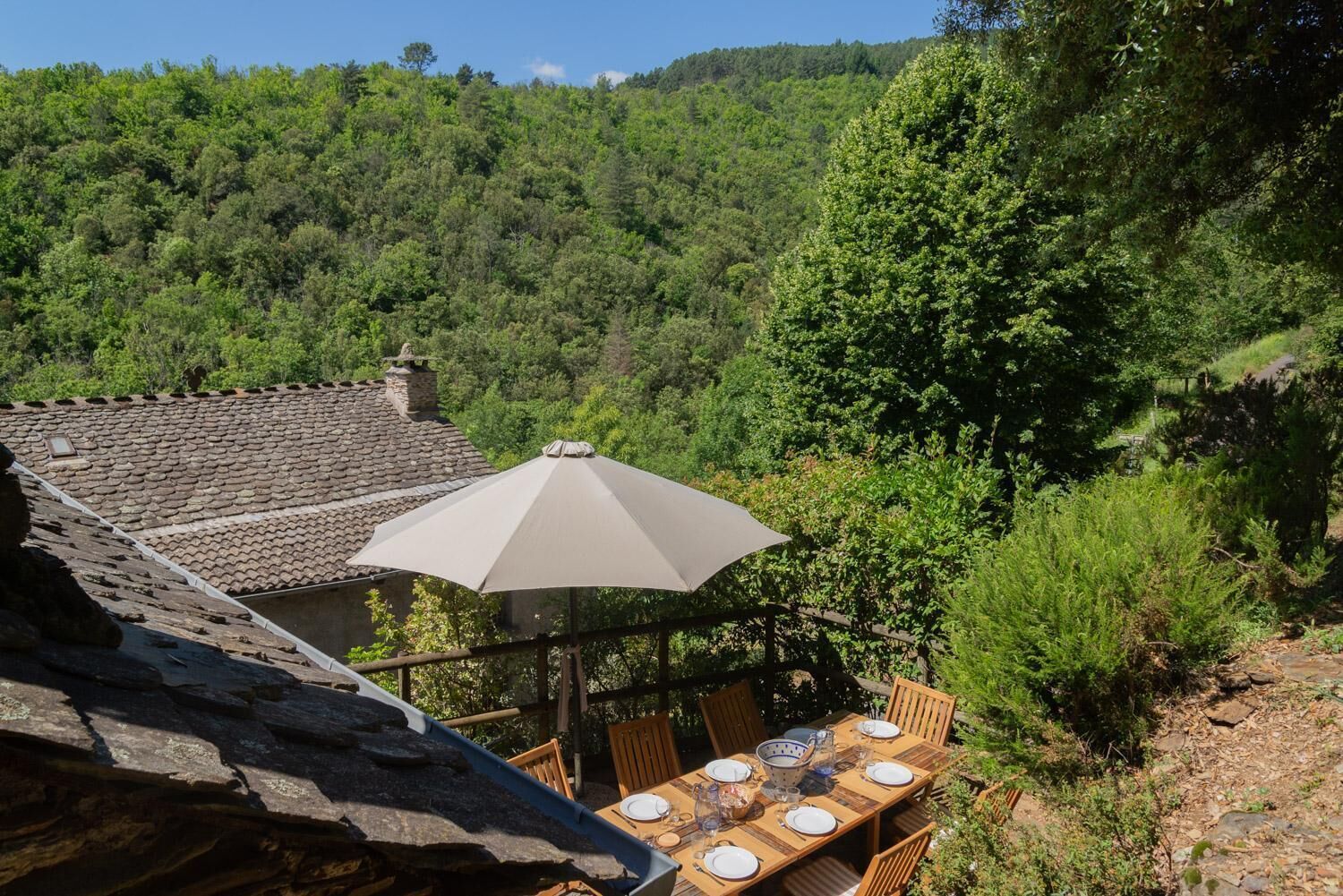 A dining area with a table set for four, an umbrella, and a view of a forested hillside.