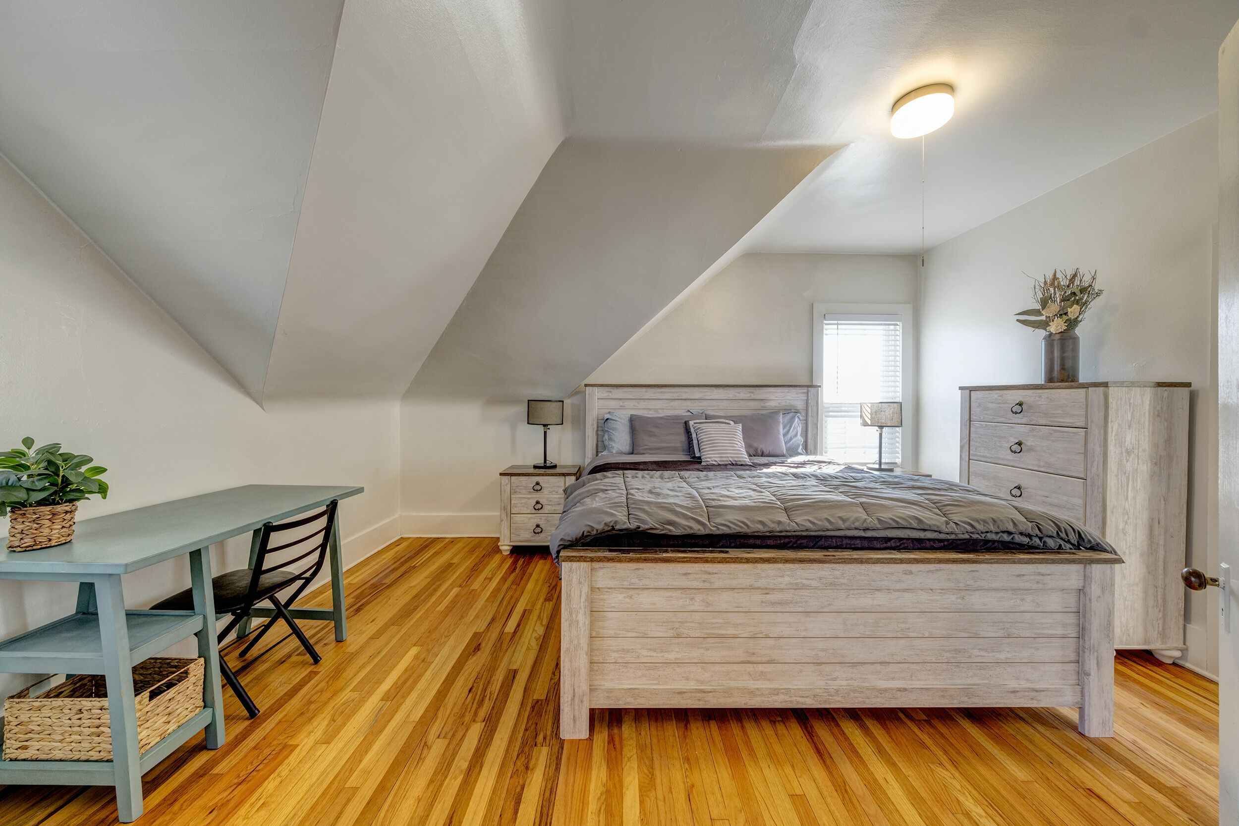 A bedroom with a sloped ceiling, a wooden bed, a desk with a chair, a nightstand, and a dresser.
