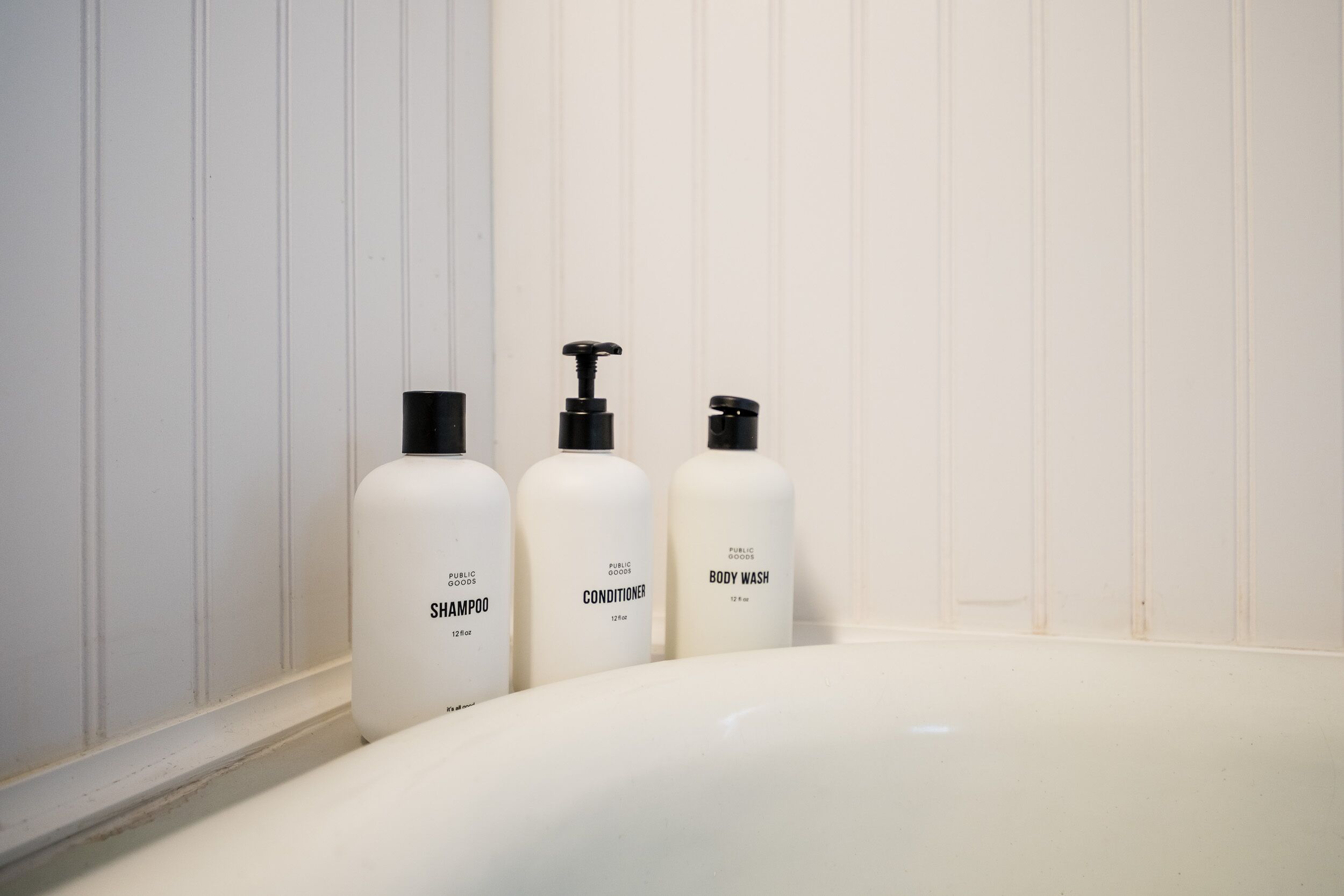 Three white bottles of public goods shampoo, conditioner, and body wash on a white sink.