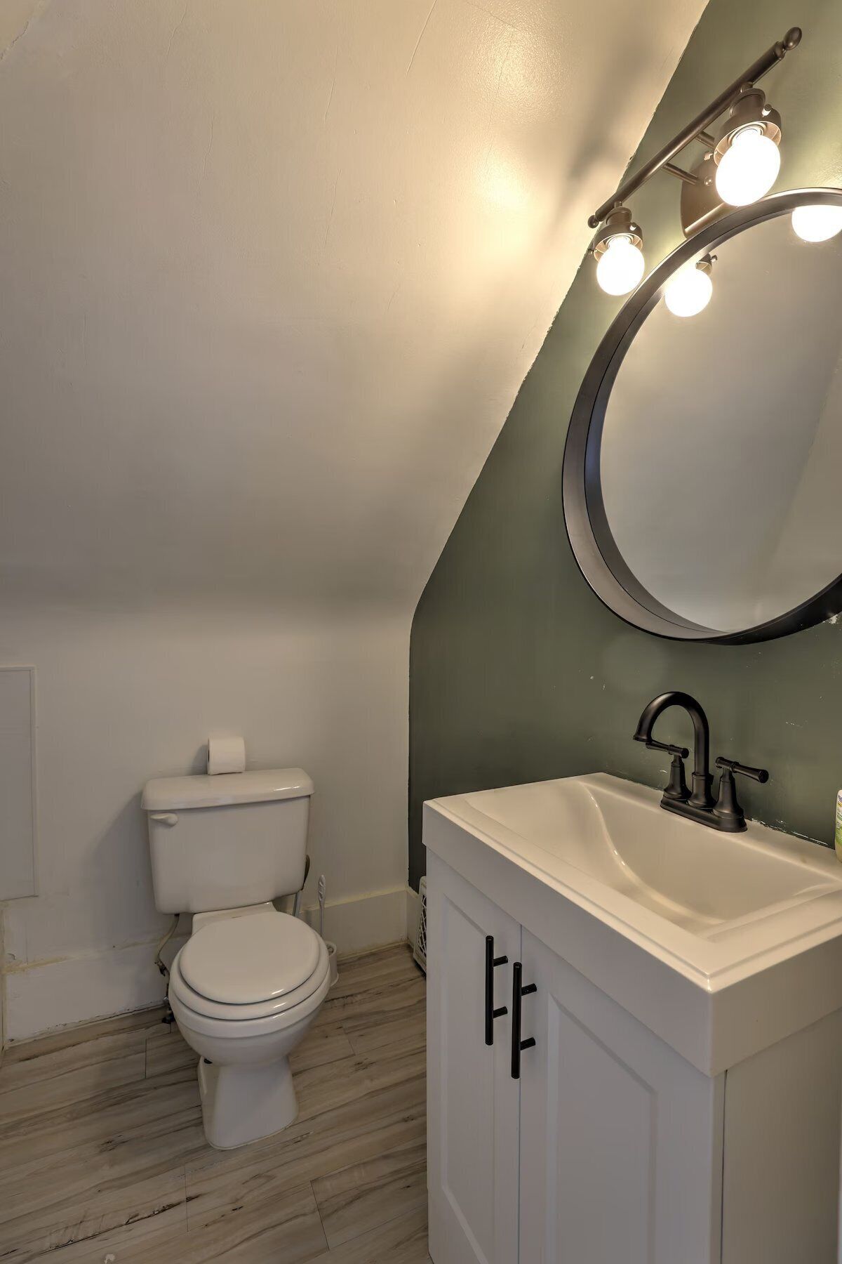 A bathroom with a white toilet, a white sink with black handles, and a mirror with lights above it.