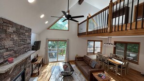 Un'ampia zona giorno con camino in pietra, ventilatore a soffitto, zona pranzo con tavolo e sedie in legno e un balcone con vista sugli alberi.