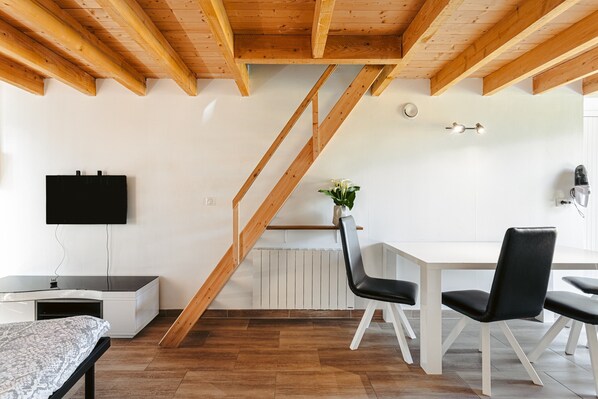 Una habitación moderna con techo de madera, una mesa blanca con sillas, un televisor negro en la pared y una escalera.