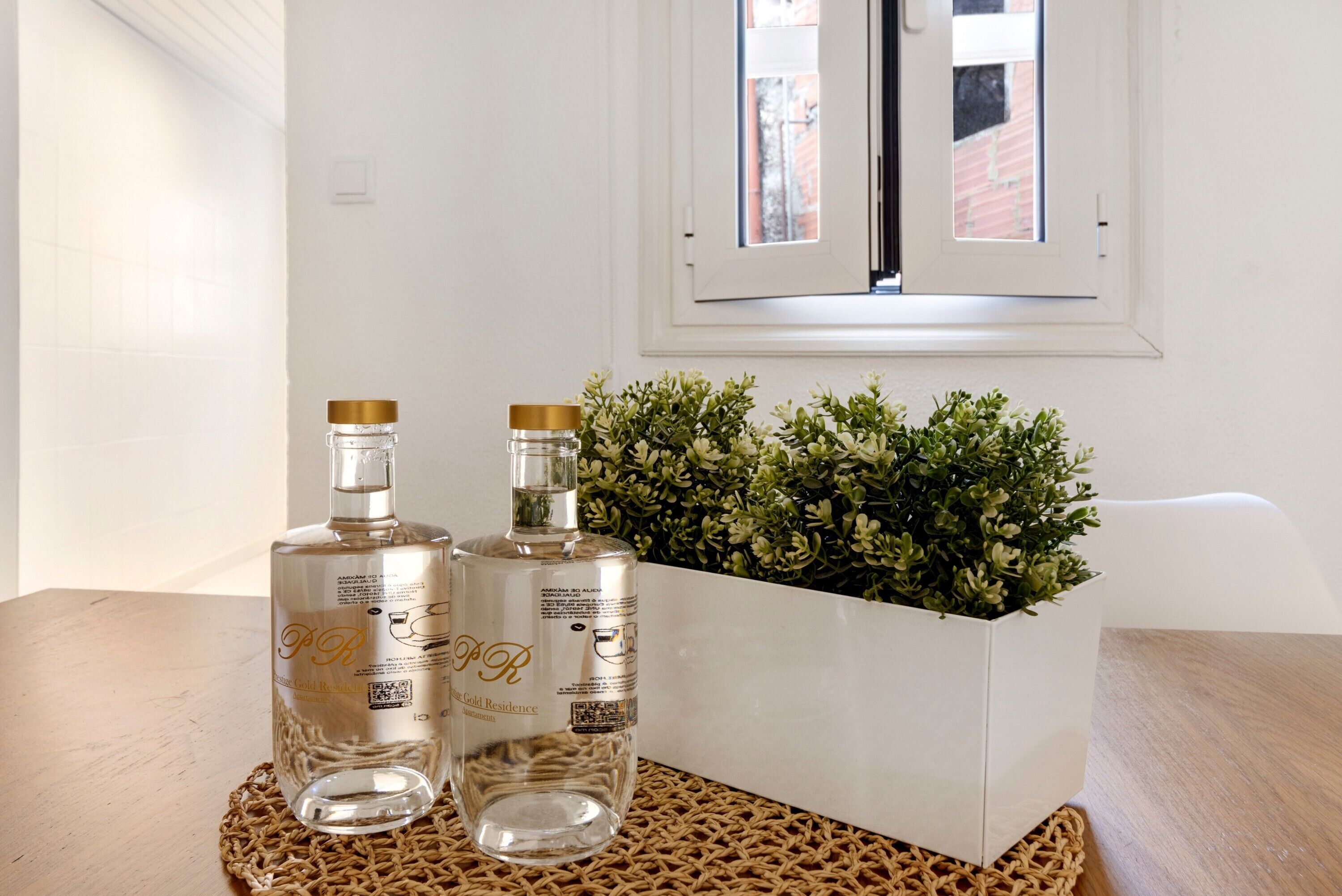 Deux bouteilles en verre transparent, coiffées de bouchons dorés, posées sur un tapis tissé, un pot de fleurs blanc contenant des plantes vertes et une fenêtre blanche donnant sur un bâtiment en briques.