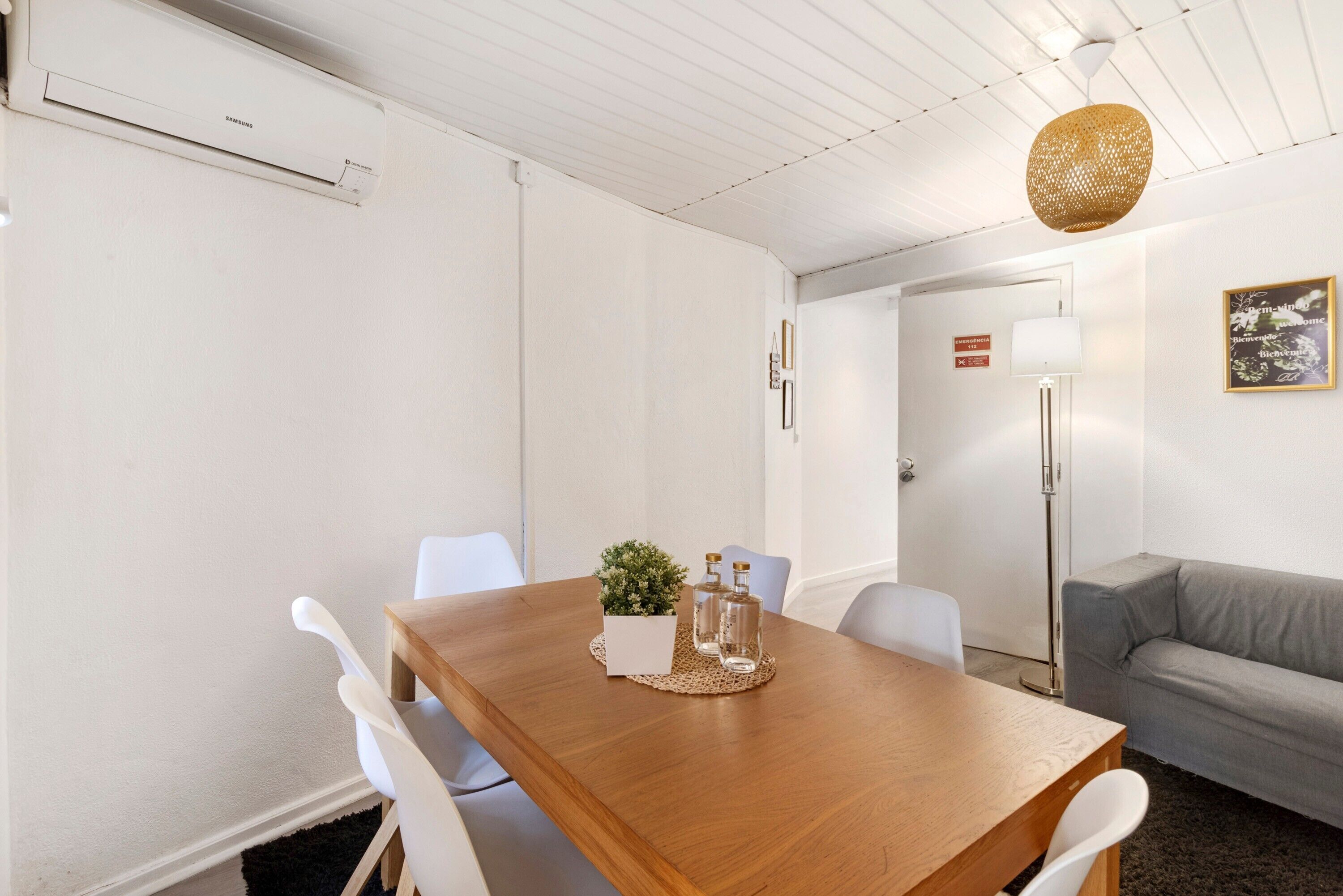 Una habitación moderna con una mesa de comedor de madera, sillas blancas, un sofá gris, una lámpara de pie y una pequeña planta en maceta.