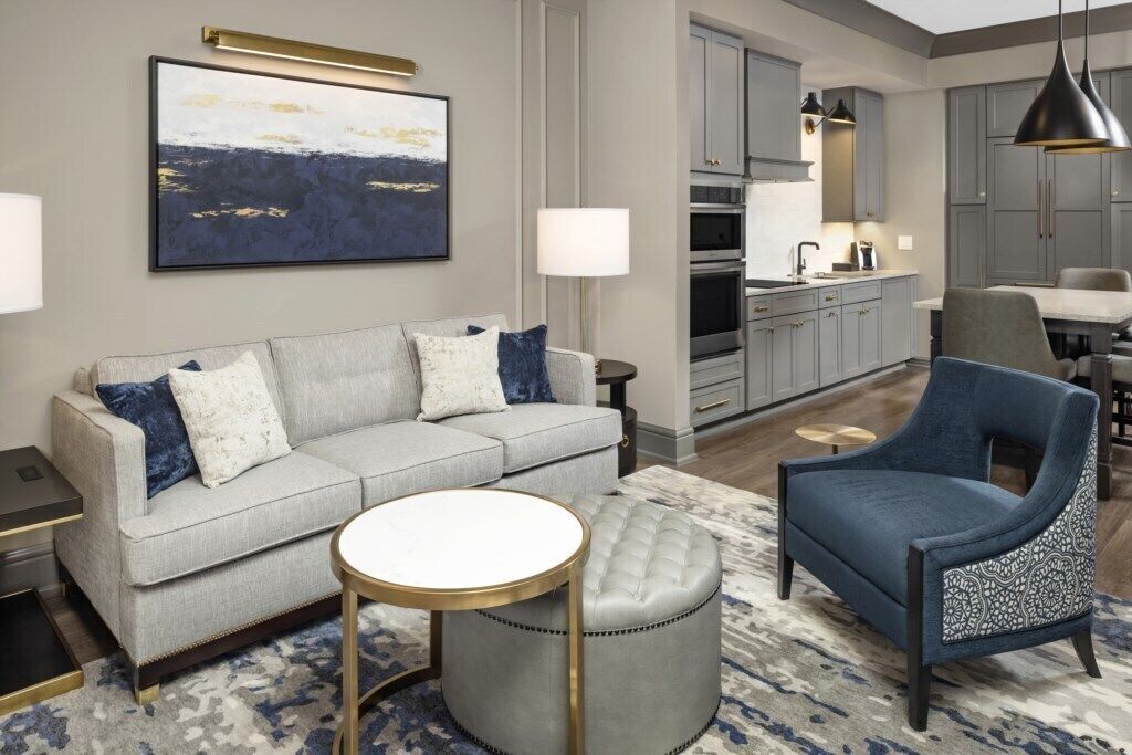 Una sala de estar moderna con un sofá gris, un sillón azul, una mesa de centro redonda y una cocina al fondo.