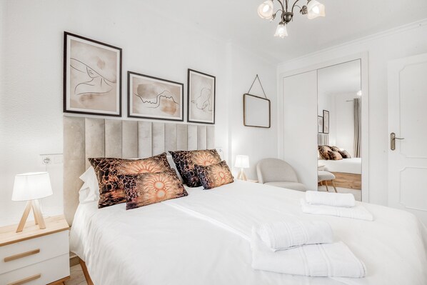 A neatly arranged bedroom with a bed, bedside tables, a lamp, framed artwork on the wall, and a mirror.