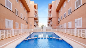 Una piscina con agua cristalina azul, rodeada por una cerca blanca, junto a un edificio con balcones y ventanas.