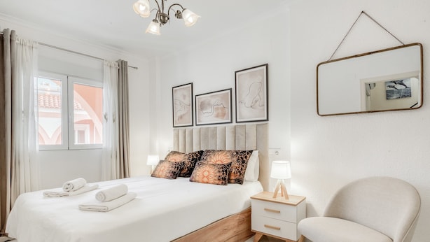 Un dormitorio con cama, mesita de noche, silla y una obra de arte en la pared.