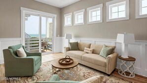 A living room with a green armchair, beige sofa, and a round coffee table. There are four windows on the wall and a balcony with a view.