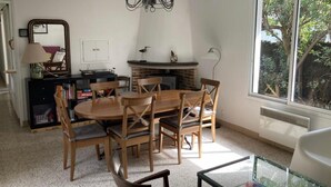 Un comedor con una mesa redonda de madera, seis sillas, una araña, una chimenea, una estantería y una ventana con vistas a árboles.