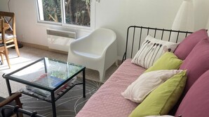 Una habitación con cama, una silla blanca, una mesa con cubierta de vidrio y una ventana con vista a árboles.
