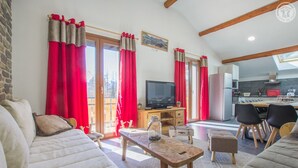 A living room with a sofa, a coffee table, a TV, and a dining area with chairs and a table. - Gîte Les Mélèzes – Mountain Views, Balcony & Wi-Fi (TERMIGNON)