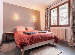 Un dormitorio con una cama, dos luces fijadas a la pared, una ventana con cortinas, un radiador y una mesita de noche de madera.