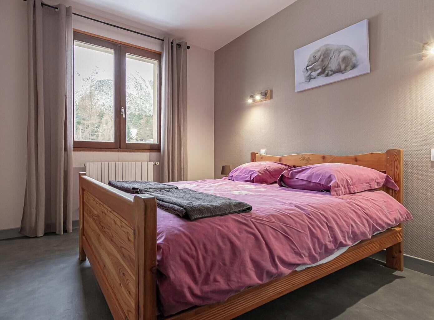 A bedroom with a wooden bed, pink bedding, a window with curtains, a radiator, and a wall-mounted light.
