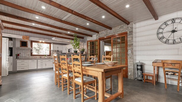 Zona pranzo con tavolo e sedie in legno, parete in pietra con orologio e cucina sullo sfondo.
