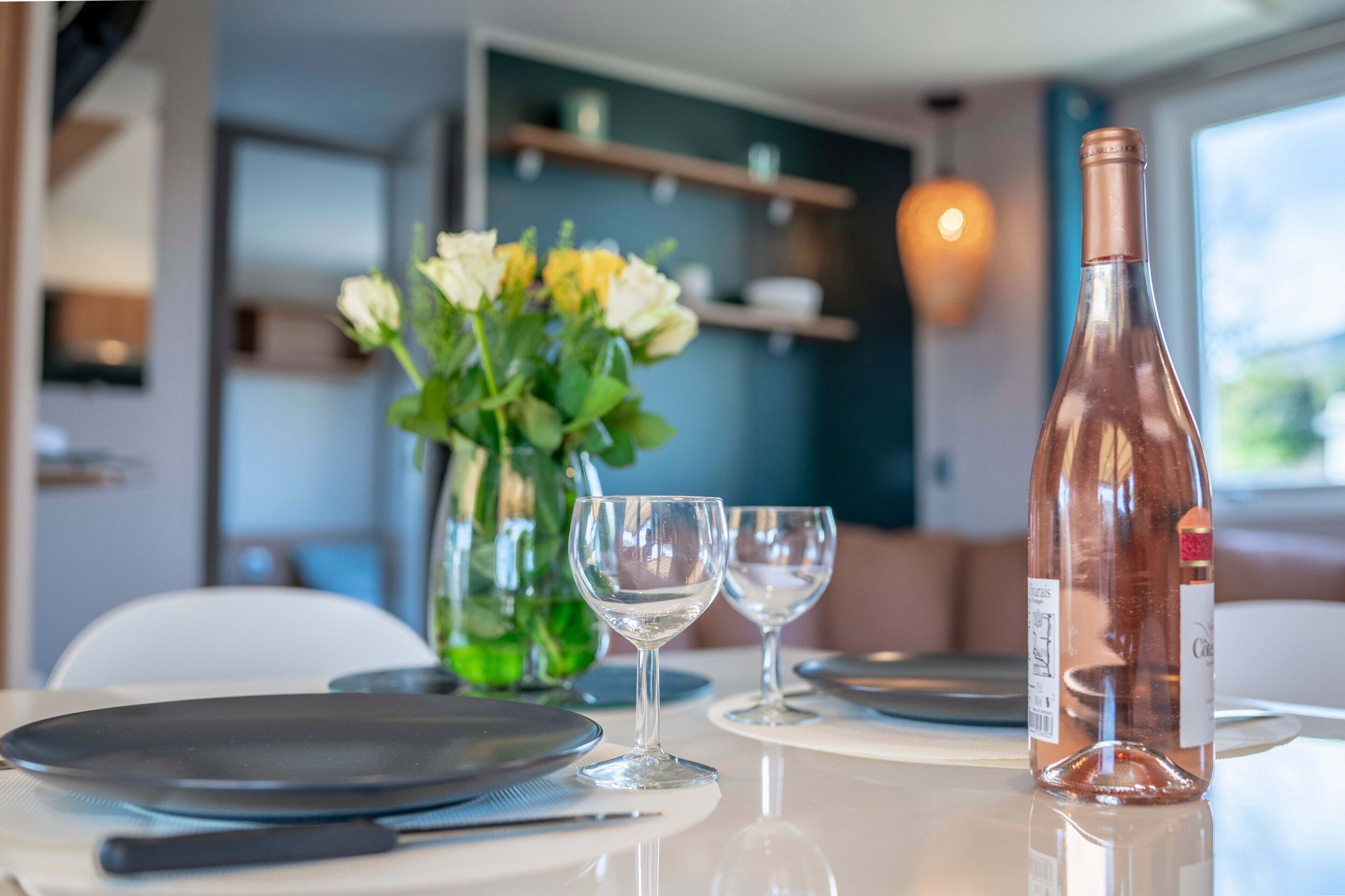 A dining table with plates, cutlery, and a bottle of wine, set in a room with a vase of flowers and a hanging light fixture.