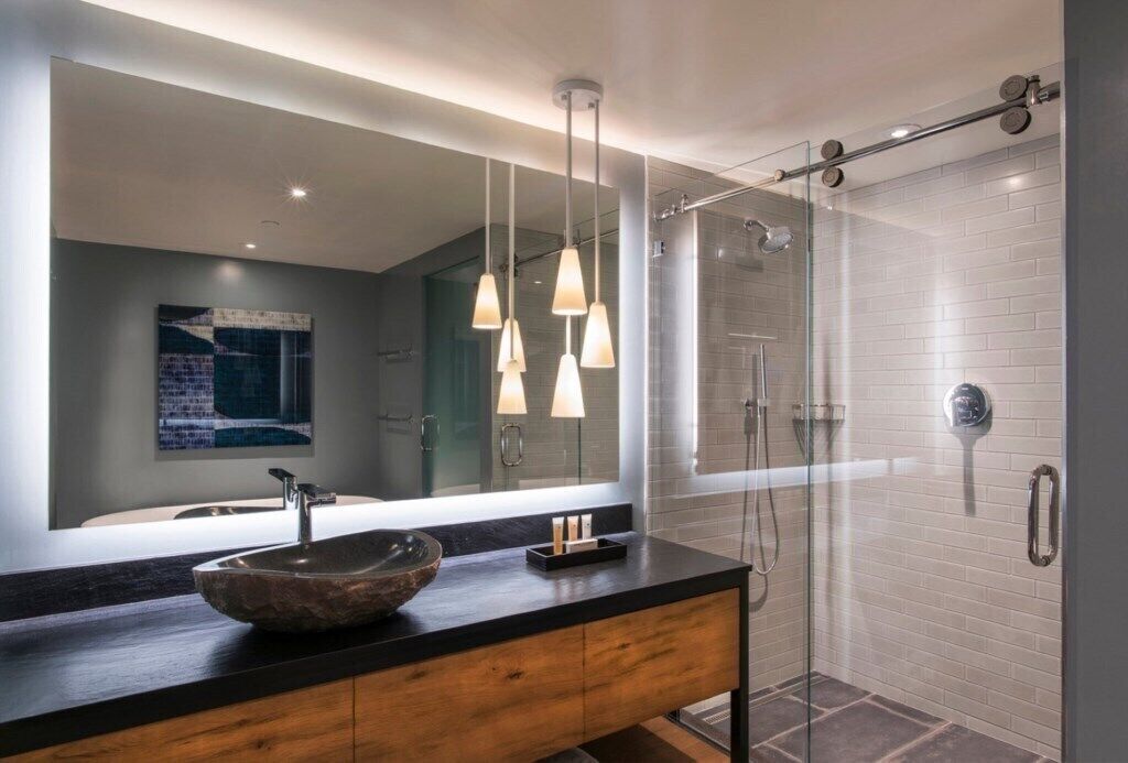 Een moderne badkamer met een grote spiegel, een houten wastafelmeubel, een stenen wasbak, hanglampen en een douche met glazen wanden.