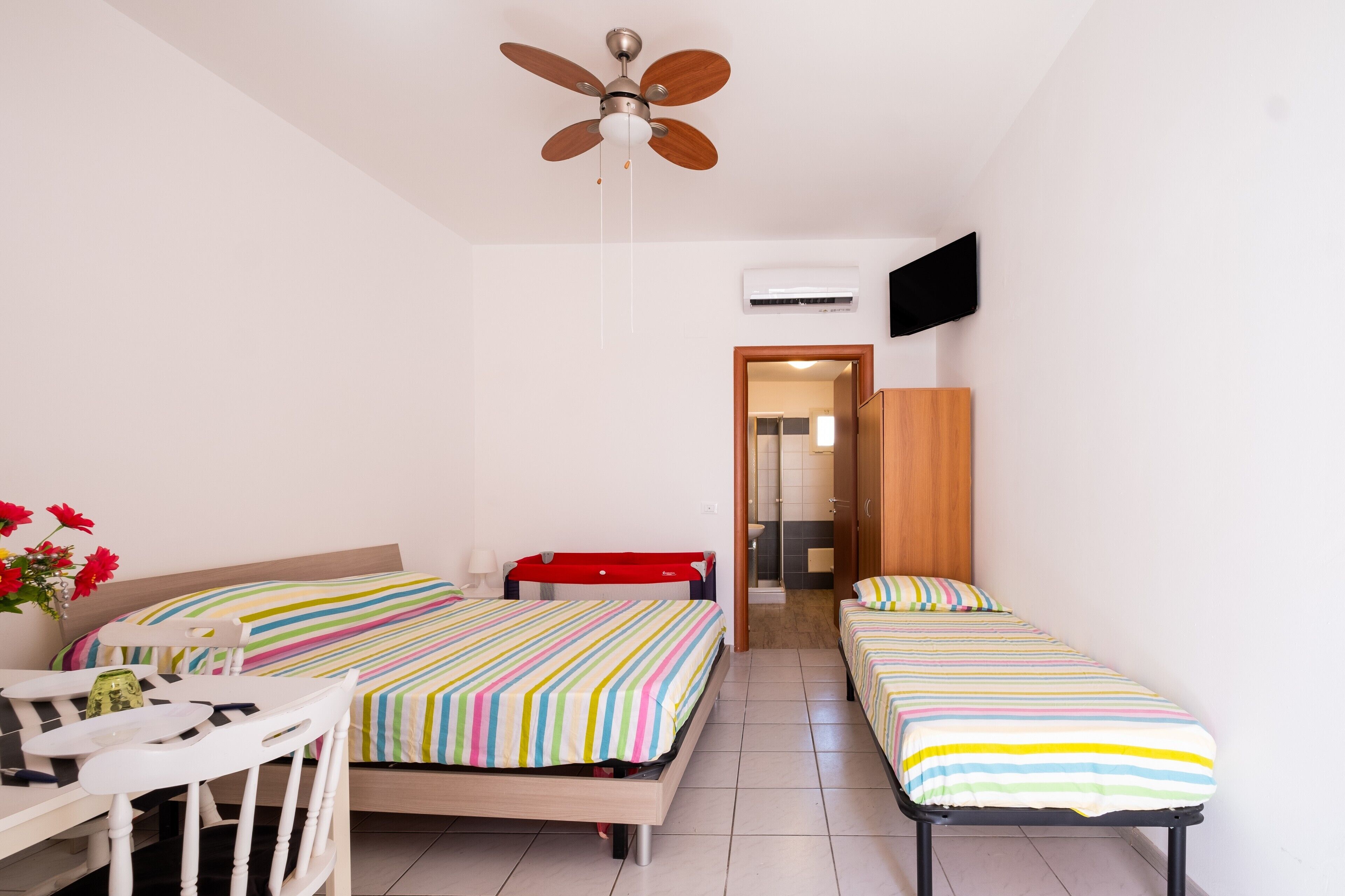A hotel room with two beds, a ceiling fan, an air conditioning unit, a TV, and a small table with chairs.