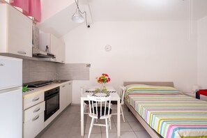 Cocina compacta con mesa y sillas, cama con colcha a rayas de colores y un reloj pequeño en la pared.