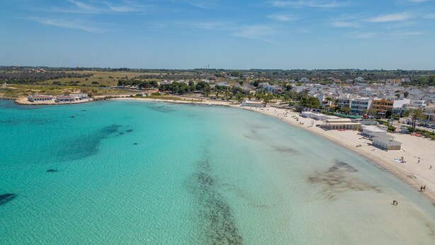Une zone côtière dotée d’une plage de sable, d’eaux turquoise et limpides, et de bâtiments qui bordent le rivage.