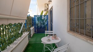 Un petit espace extérieur avec une table et des chaises blanches, une porte bleue et une fenêtre ornée de barreaux en métal.