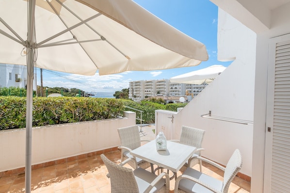 A balcony with a table and chairs, a white umbrella, and a view of buildings and greenery.
