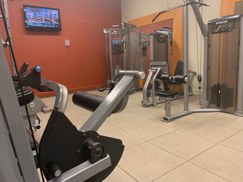 Une salle de sport équipée de divers appareils d’exercice, avec un téléviseur fixé au mur et une porte donnant sur une autre pièce.