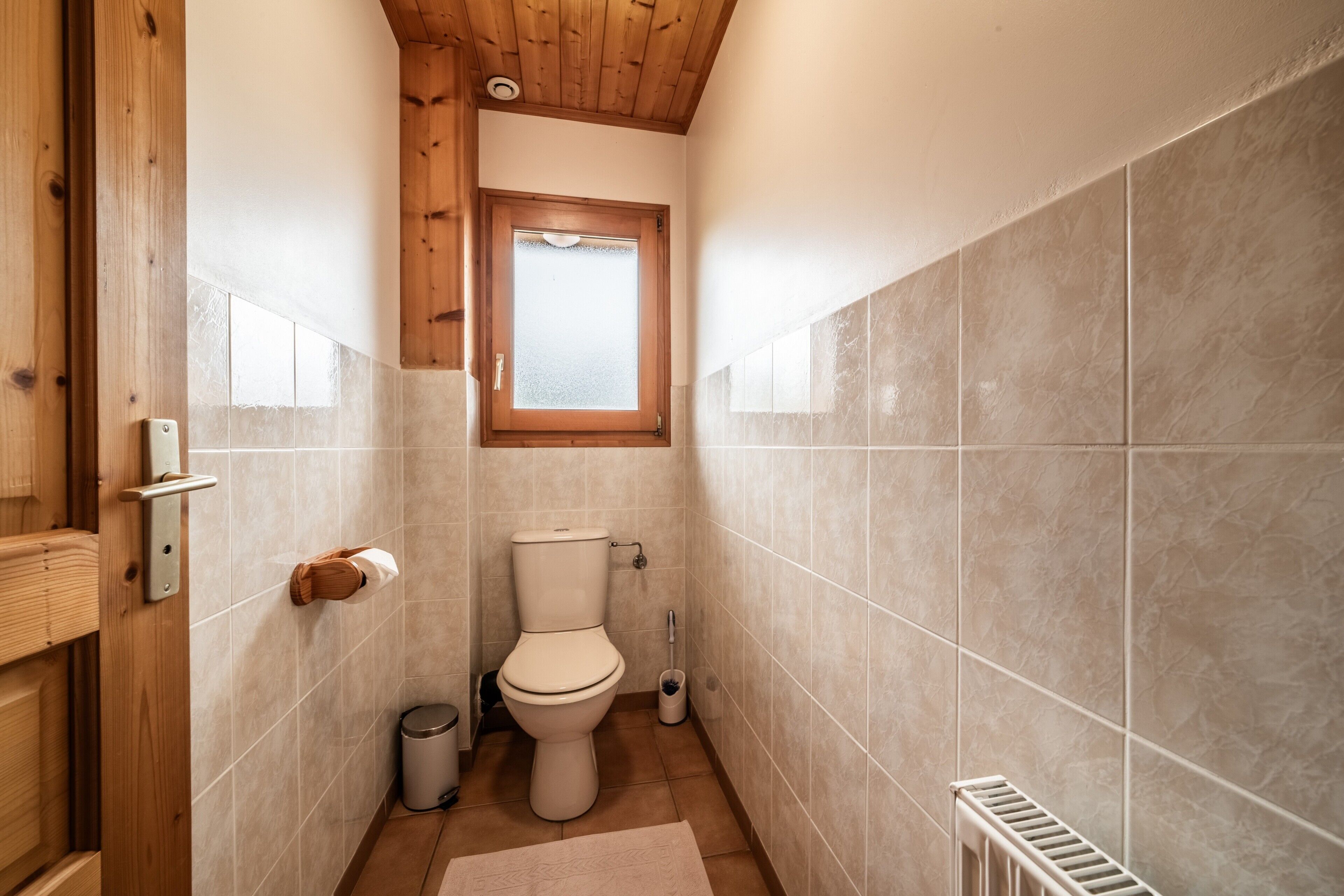A compact bathroom with a toilet, a window, a radiator, and a wooden door.