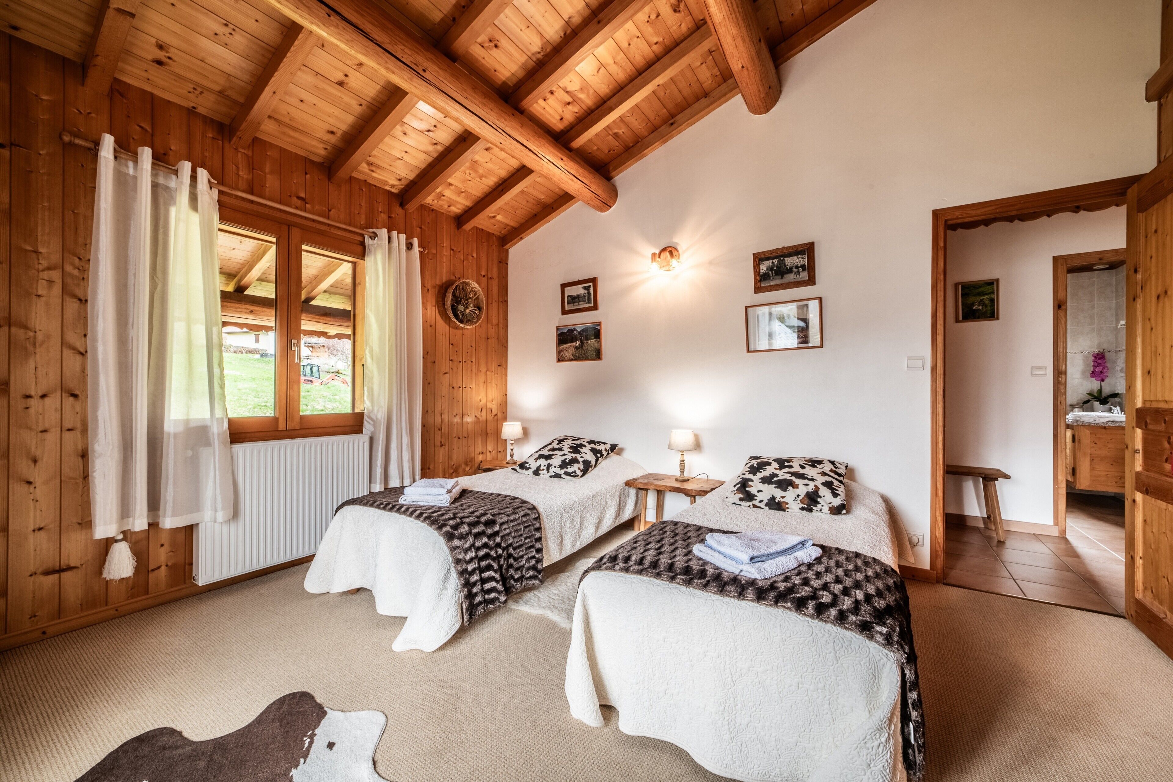 A wooden cabin bedroom with two beds, a window with curtains, a wall clock, and framed pictures.